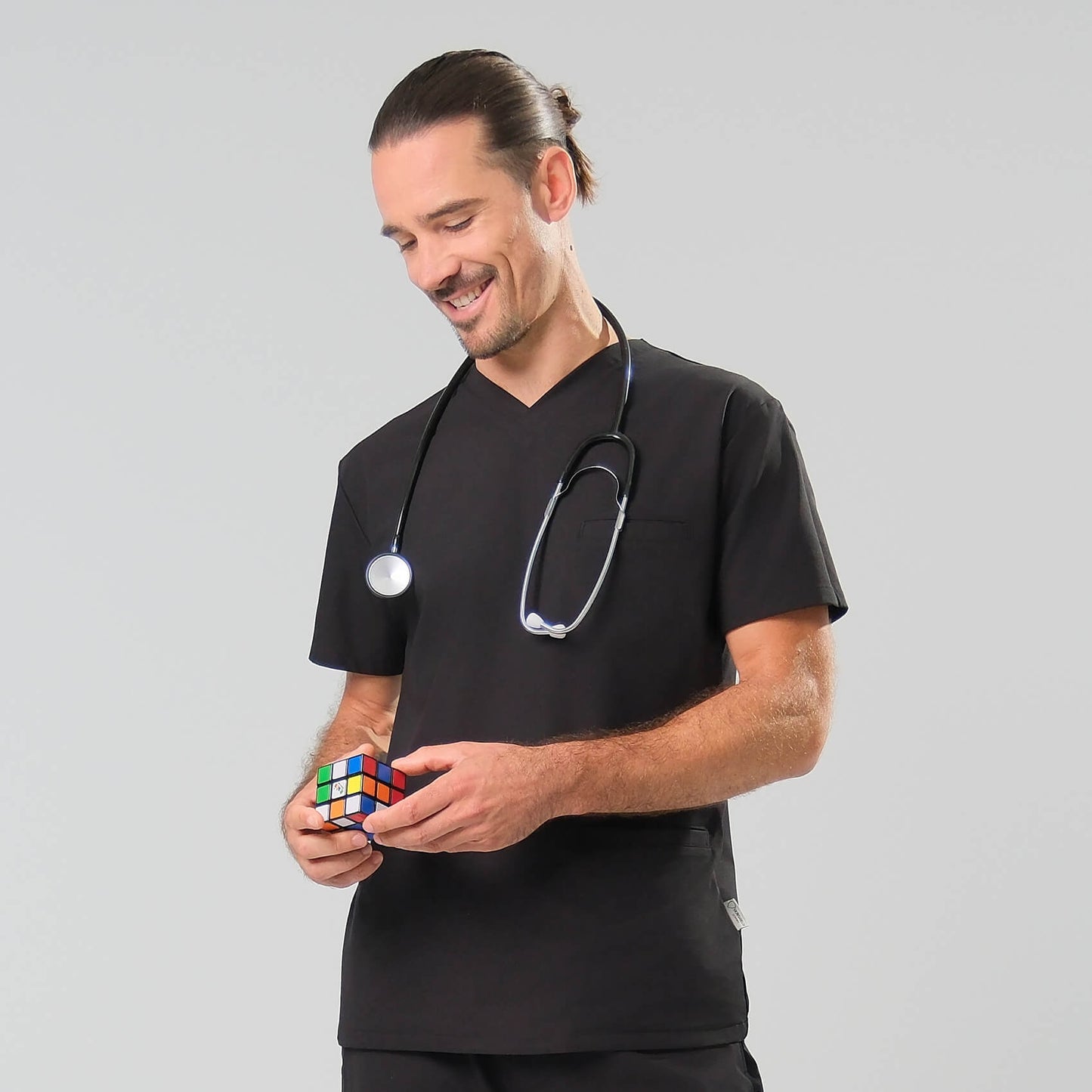 Man in black scrubs with a stethoscope around his neck holding a Rubik's Cube against a grey background