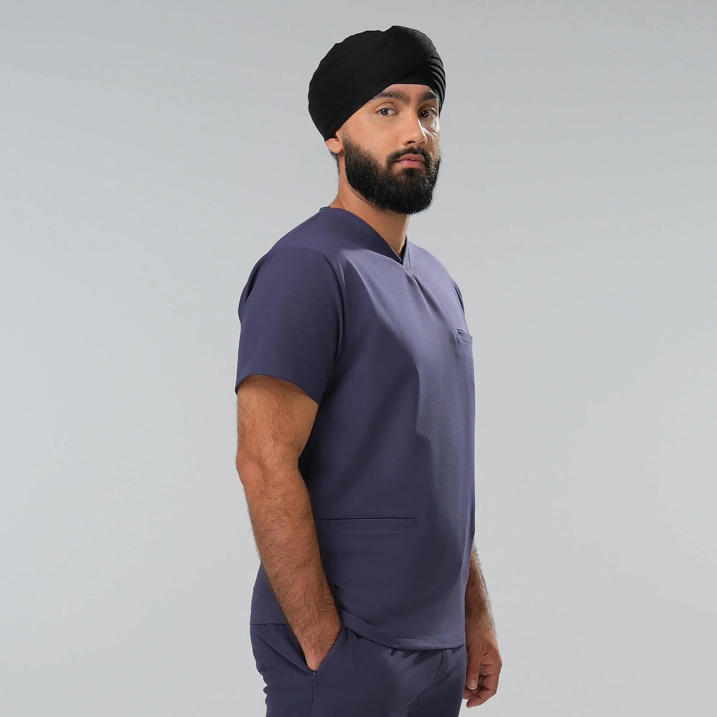 Man wearing a navy blue scrub top and trousers with a black turban on a grey background