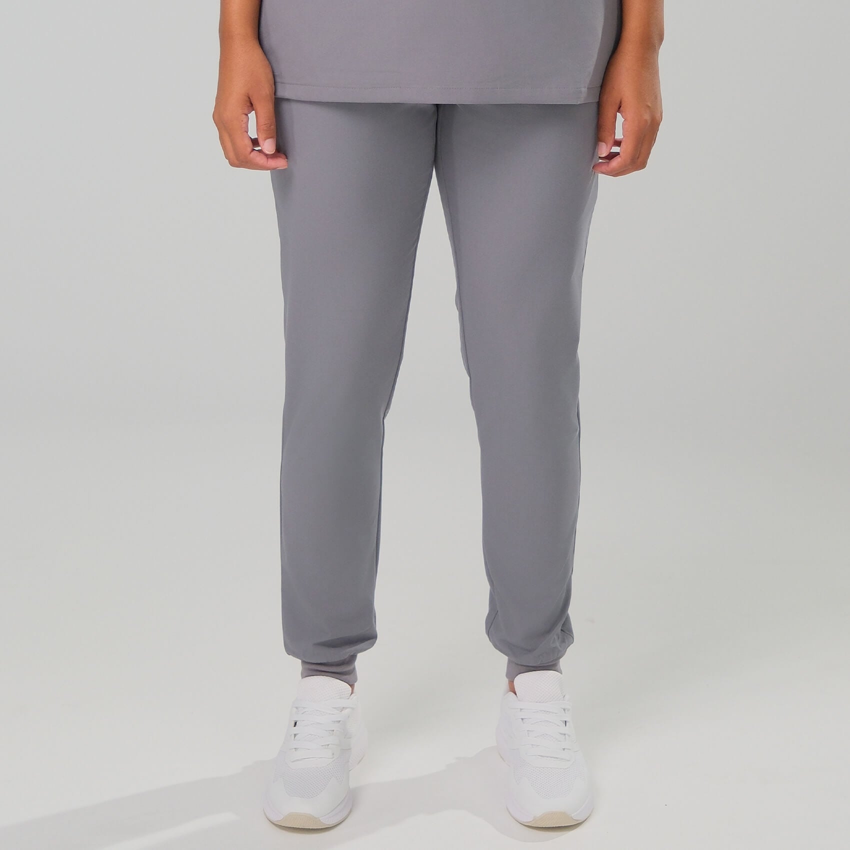 Woman wearing grey tapered scrub joggers and white trainers on a plain background